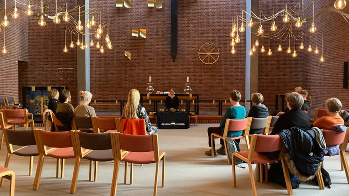 Mennesker siddende i et rummeligt, veloplyst lokale med moderne belysning og et centralt sceneområde.