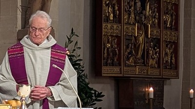 Ein Priester in liturgischer Kleidung führt eine religiöse Zeremonie mit Kerzen und religiösen Kunstwerken im Hintergrund durch.