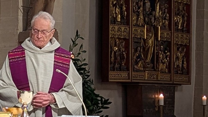 Ein Priester in liturgischer Kleidung führt eine religiöse Zeremonie mit Kerzen und religiösen Kunstwerken im Hintergrund durch.