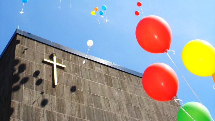 Bunte Luftballons schweben über einem Gebäude mit einem Kreuzsymbol an der Fassade unter einem klaren blauen Himmel.