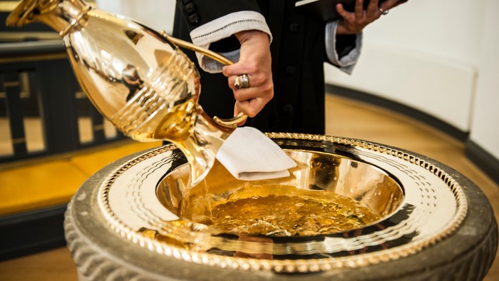 En person hælder hellig olie fra et gyldent kar ned i en stor beholder under en religiøs velsignelsesceremoni.
