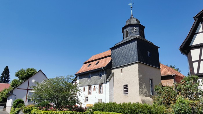 Ein weißes Gebäude mit schwarzem Dach und Uhrturm, umgeben von grünen Büschen und Bäumen