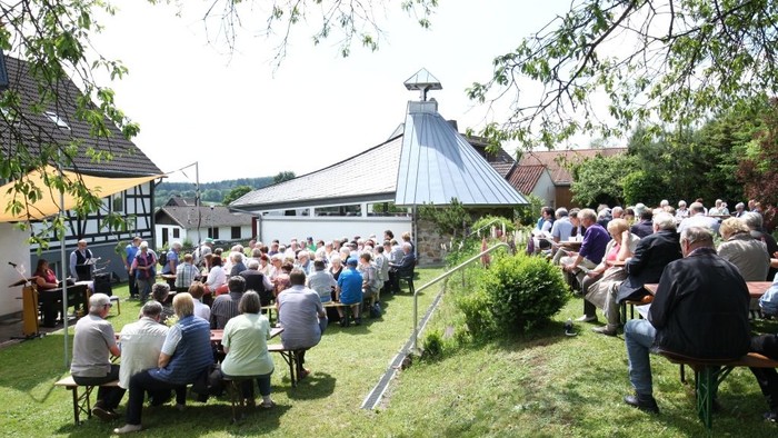 Große Menschenmenge sitzt auf Stühlen im Freien, umgeben von Bäumen und Gebäuden.
