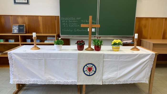 Ein einfacher Altaraufbau in einem Klassenzimmer mit Blumen, Kerzen und einem religiösen Symbol auf einem weißen Tuch.