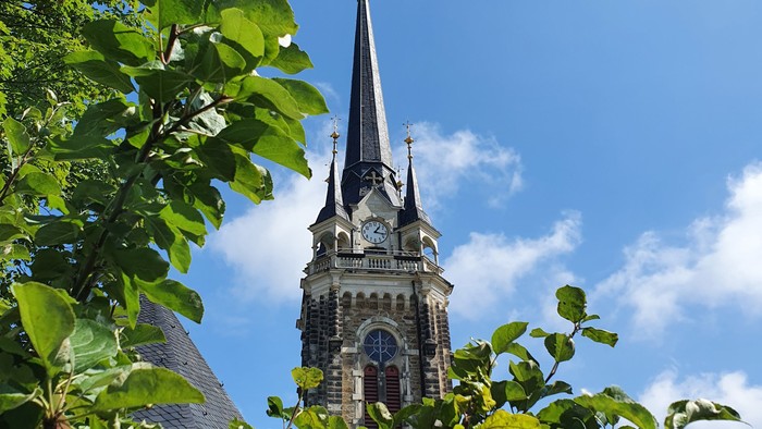 Ein hoher Kirchturm mit einer Uhr erhebt sich über üppiges grünes Laub unter einem klaren blauen Himmel.