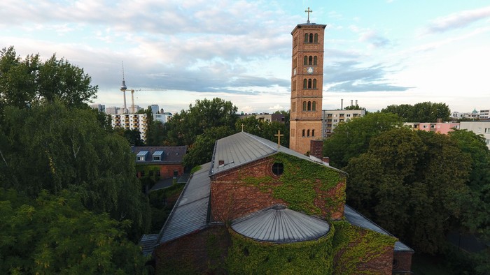 Historische Backsteinkirche, von Efeu bewachsen, umgeben von üppigem Grün und einer städtischen Skyline im Hintergrund.