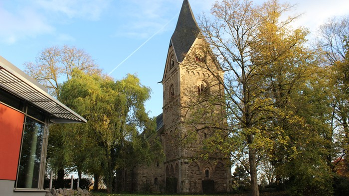 Altes Steingebäude mit spitzem Turm neben Bäumen und rotem Haus