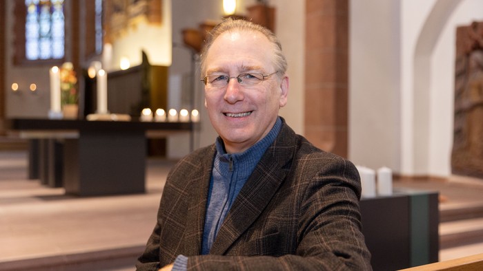 Ein lächelnder Mann im Anzug sitzt in einer Kirchenbank, trägt eine Brille und eine karierte Jacke.