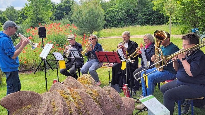 Kleine Bläsergruppe spielt im Freien: Mehrere Musikerinnen und Musiker sitzen mit Trompeten, Posaune und Tuba auf Stühlen und musizieren nach Notenständern, während links ein Mann im Stehen dirigiert oder selbst Trompete spielt. Die Szene spielt auf einer