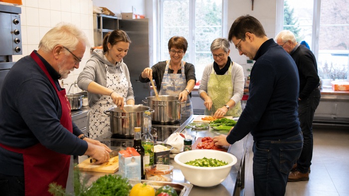 Mehrere Menschen unterschiedlichen Alters stehen gemeinsam in der hellen Küche eines Gemeindehauses um eine große Arbeitsfläche. Sie schneiden Gemüse, rühren in Töpfen und tauschen sich dabei lebhaft aus.