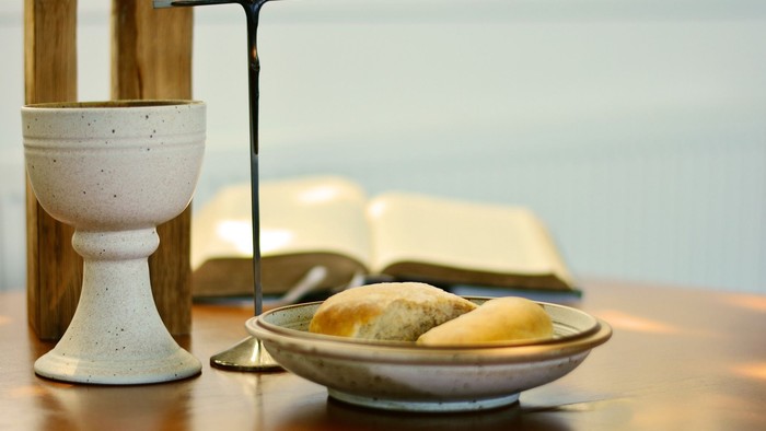 Ein Kelch, ein Kreuz und ein offenes Buch liegen auf einem Tisch neben einem Teller mit Brot.