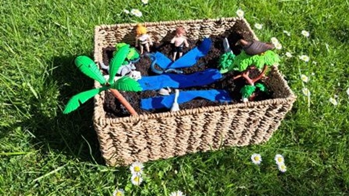 Ein Weidenkorb, gefüllt mit Spielzeugpflanzen und Erde, umgeben von kleinen weißen Blumen auf Gras.