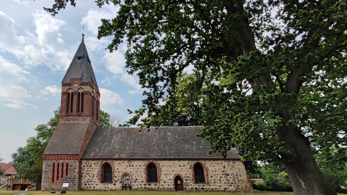 Das Bild zeigt eine alte Kirche mit hohem Turm und Steinmauern, umgeben von Bäumen und grünem Gras.