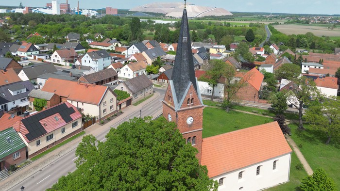 Luftaufnahme eines malerischen Dorfes mit einer markanten Kirche mit hohem Kirchturm und umliegenden Häusern mit roten Dächern.