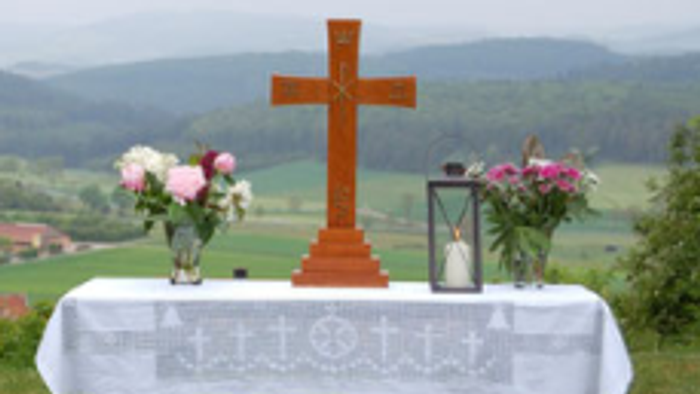 Das Foto zeigt einen aufgebauten Altar auf einer Wiese. Der Tisch trägt ein Kreuz, 2 Blumensträuße links und rechts und eine Kerze.