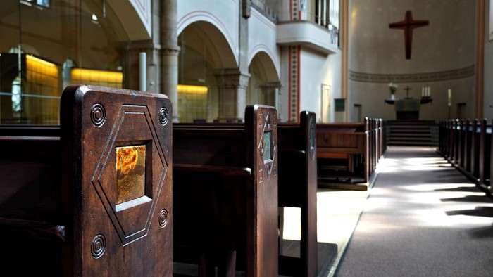 Ein ruhiger Kircheninnenraum mit hölzernen Kirchenbänken, gewölbten Säulen und einem Kreuz, das von sanftem Licht erleuchtet wird.