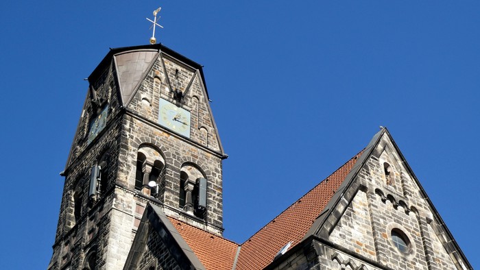 Historische Steinkirche mit hohem Glockenturm und steilem, rotziegelgedecktem Dach vor klarem blauem Himmel.