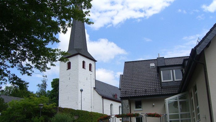Ein rotes Auto steht vor einem weißen Kirchturm und einem modernen Haus.
