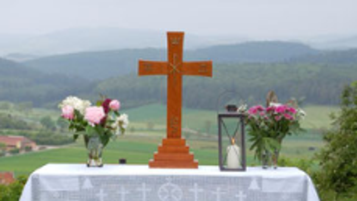 Das Foto zeigt einen aufgebauten Altar auf einer Wiese. Der Tisch trägt ein Kreuz, 2 Blumensträuße links und rechts und eine Kerze.