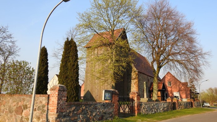 Ein rotes Backsteingebäude mit spitzem Dach und hohem Tor, umgeben von Bäumen und einem Laternenpfahl.