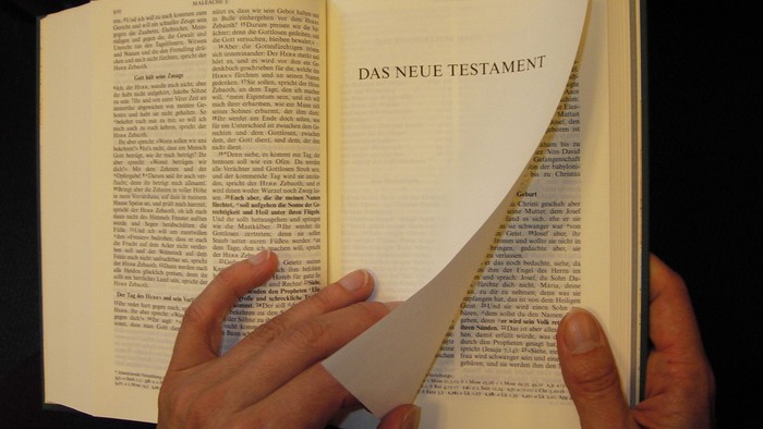 Hände halten ein offenes Buch, auf dem die Seite *„Das Neue Testament“* zu sehen ist.