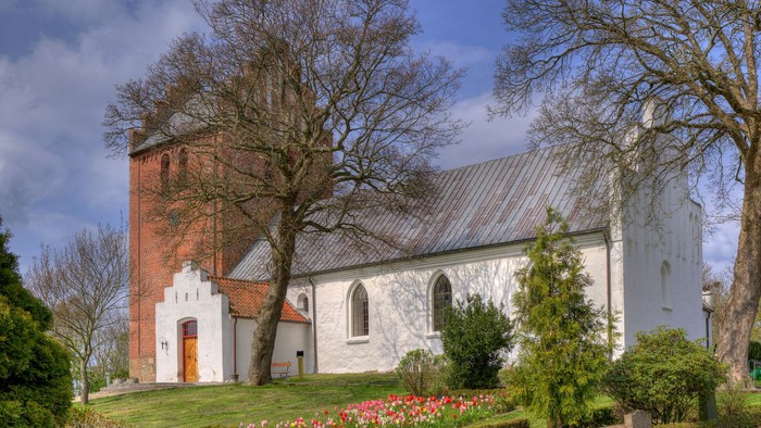 Hyggelig kirke med rødt murstens tårn og hvide mure, gemt i en fredfyldt, anlagt have.
