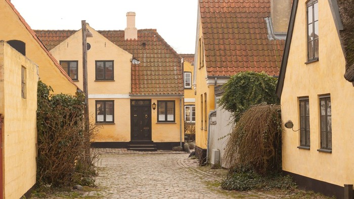 Hyggelig brostensbelagt gyde omkranset af charmerende gule huse og frodig grønne beplantning.