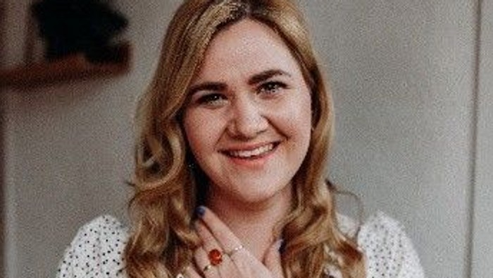 Eine fröhliche Frau in einem weißen Kleid mit Punkten posiert selbstbewusst drinnen mit einem warmen Lächeln.