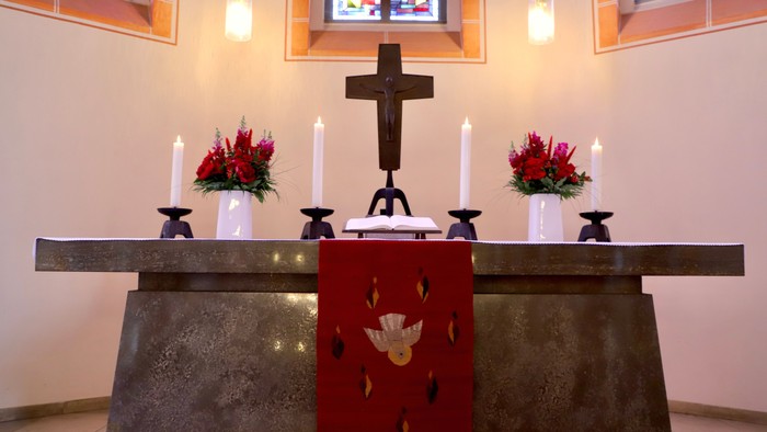 Eleganter Altar mit einem Kreuz, Kerzen, Blumengestecken und einem roten Tuch mit einem Taubenemblem.