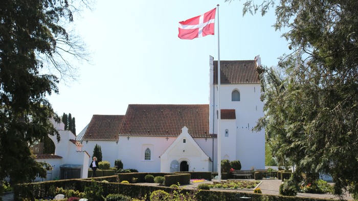 En idyllisk hvid kirke med rødt tag og et dansk flag vajende på toppen af tårnet, omgivet af frodig grøn natur.