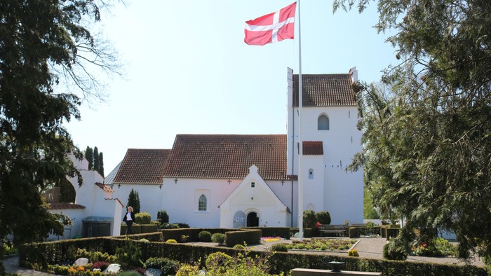 En idyllisk hvid kirke med rødt tag og et dansk flag vajende på toppen af tårnet, omgivet af frodig grøn natur.