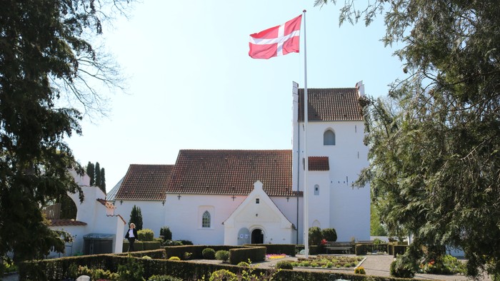 En idyllisk hvid kirke med rødt tag og et dansk flag vajende på toppen af tårnet, omgivet af frodig grøn natur.