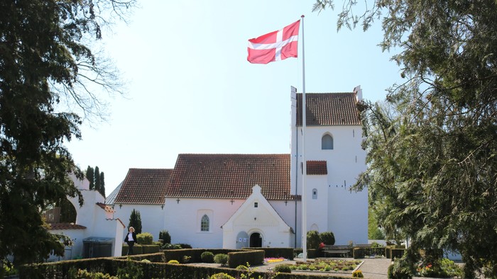 En idyllisk hvid kirke med rødt tag og et dansk flag vajende på toppen af tårnet, omgivet af frodig grøn natur.