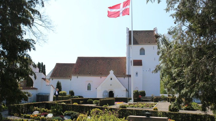 En idyllisk hvid kirke med rødt tag og et dansk flag vajende på toppen af tårnet, omgivet af frodig grøn natur.