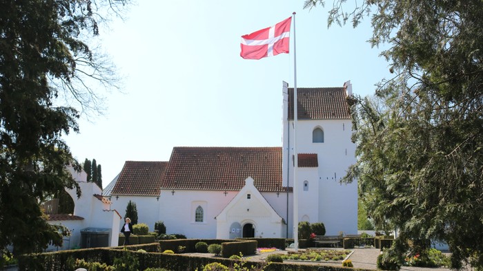 En idyllisk hvid kirke med rødt tag og et dansk flag vajende på toppen af tårnet, omgivet af frodig grøn natur.