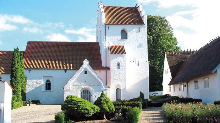 En smuk og hvid kirke med tårn og røde tag i en trædit og grønne omgivelser