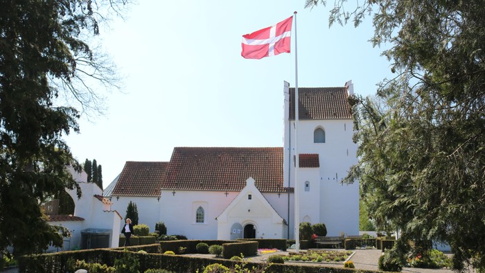 En idyllisk hvid kirke med rødt tag og et dansk flag vajende på toppen af tårnet, omgivet af frodig grøn natur.