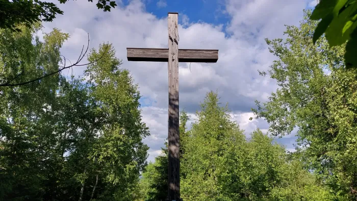 Ein hölzernes Kreuz steht hoch vor grünem Laub und blauem Himmel.