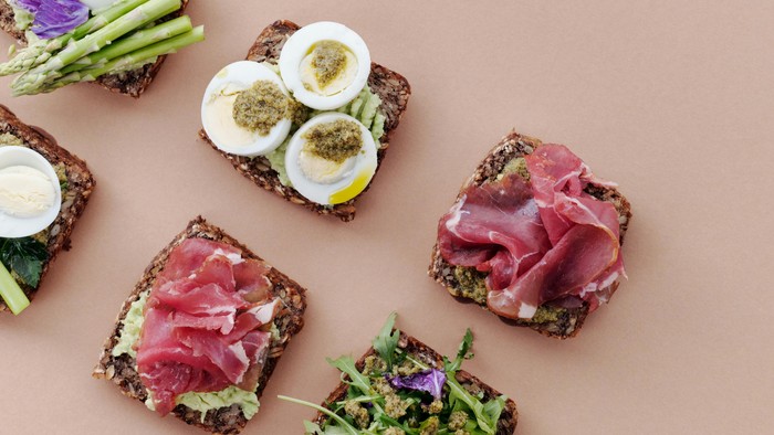 Farverigt åbent smørrebrød med æg, laks, grøntsager, asparges og urter på en blød baggrund.