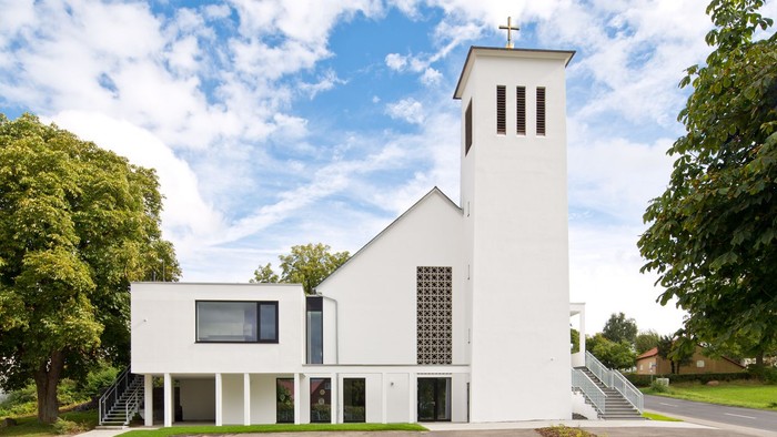 Moderne Kirche mit schlankem weißem Design, hohem Glockenturm und umgeben von üppigem Grün unter blauem Himmel.