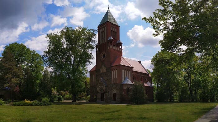 Ein Kirchengebäude mit hohem Turm steht auf einem grünen Hügel, umgeben von Bäumen und Häusern.