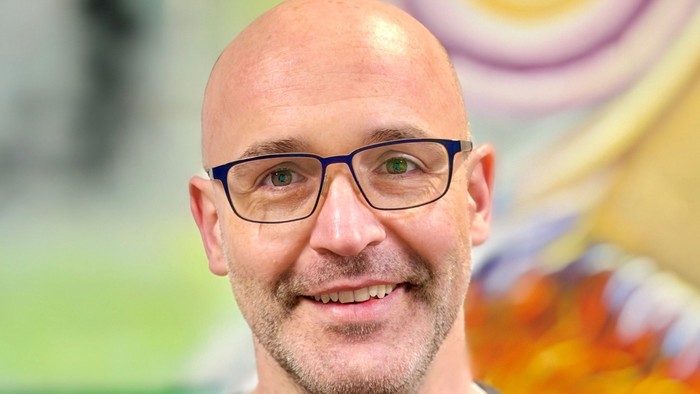 En smilende skaldet mand med briller står foran en farverig, abstrakt baggrund.