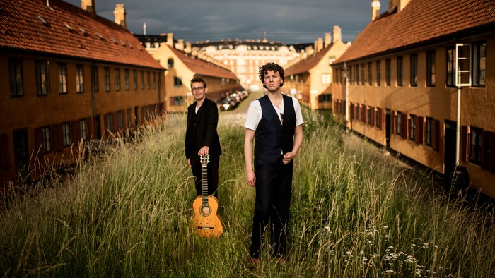 To musikere står i højt græs mellem historiske gule murstensbygninger med en guitar og poser sammen.
