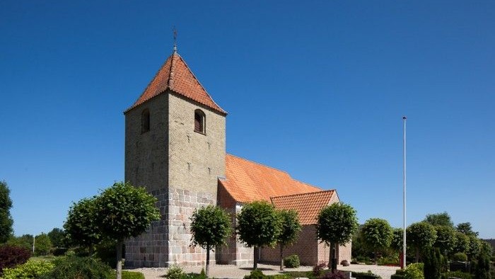 En lille kirke med tårn og røde tag, omgivet af træer og buske under et klarblåt sky.