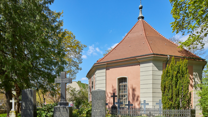 Ein kleiner rosa und weißer Glockenturm mit spitzem Dach in einem Friedhof, umgeben von Bäumen und Grabsteinen.