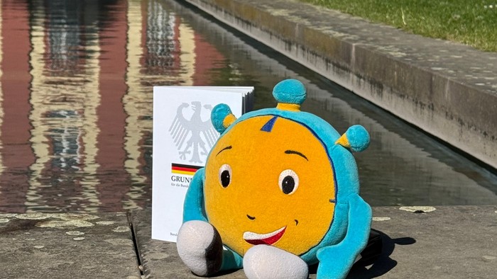 Das Maskottchen Stoppi der 72-Stunden-Aktion, ein rundes, gelb-blaues Plüschtier mit lachendem Gesicht und weißen Händen, sitzt auf einer Steinmauer am Wasserbecken im Dechaneigarten beim Fuldaer Dom. Neben Stoppi steht das Grundgesetz. Im Hintergrund sin