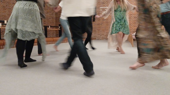 Mennesker, der danser i en cirkel, nogle barfodede, i et lokale med murstensvægge.