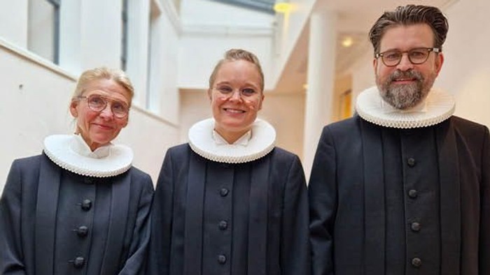 Tre personer i formelle kjoler står ved siden af en stor bikage, der tappes, med honning, der drypper ned i beholdere.