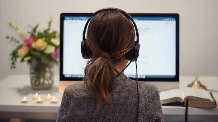 Eine Frau von hinten mit einem Headset. Sie sitzt vor dem Monitor. Auf der linken Seite sieht man Blumen und Kerzen auf dem Tisch. Auf der rechten Seite eine Bibel.