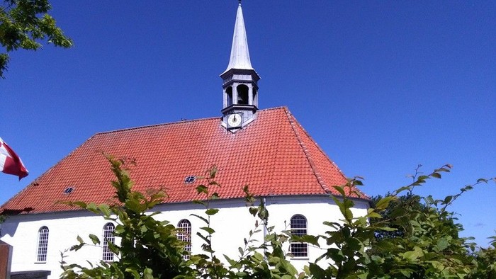En charmerende hvid kirke med rødt tag og spir omgivet af frodigt grønt løv under en klar blå himmel.